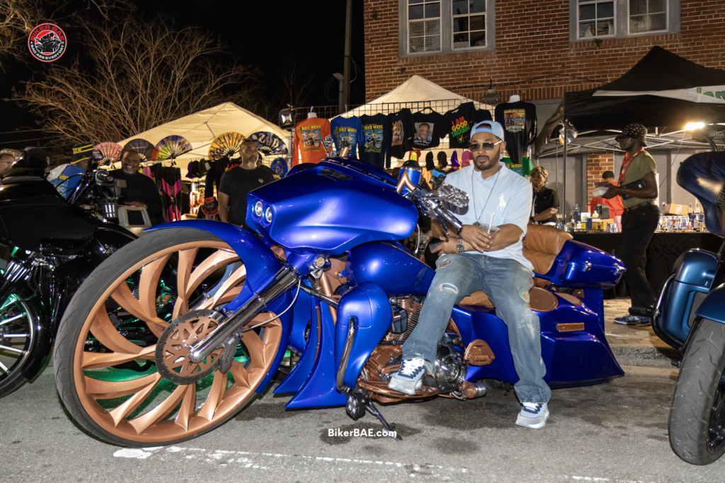 Custom blue bagger motorcycle with oversized bronze front wheel parked at Black Bike Week in Daytona Beach while a rider sits on the bike surrounded by vendors and crowds at night.